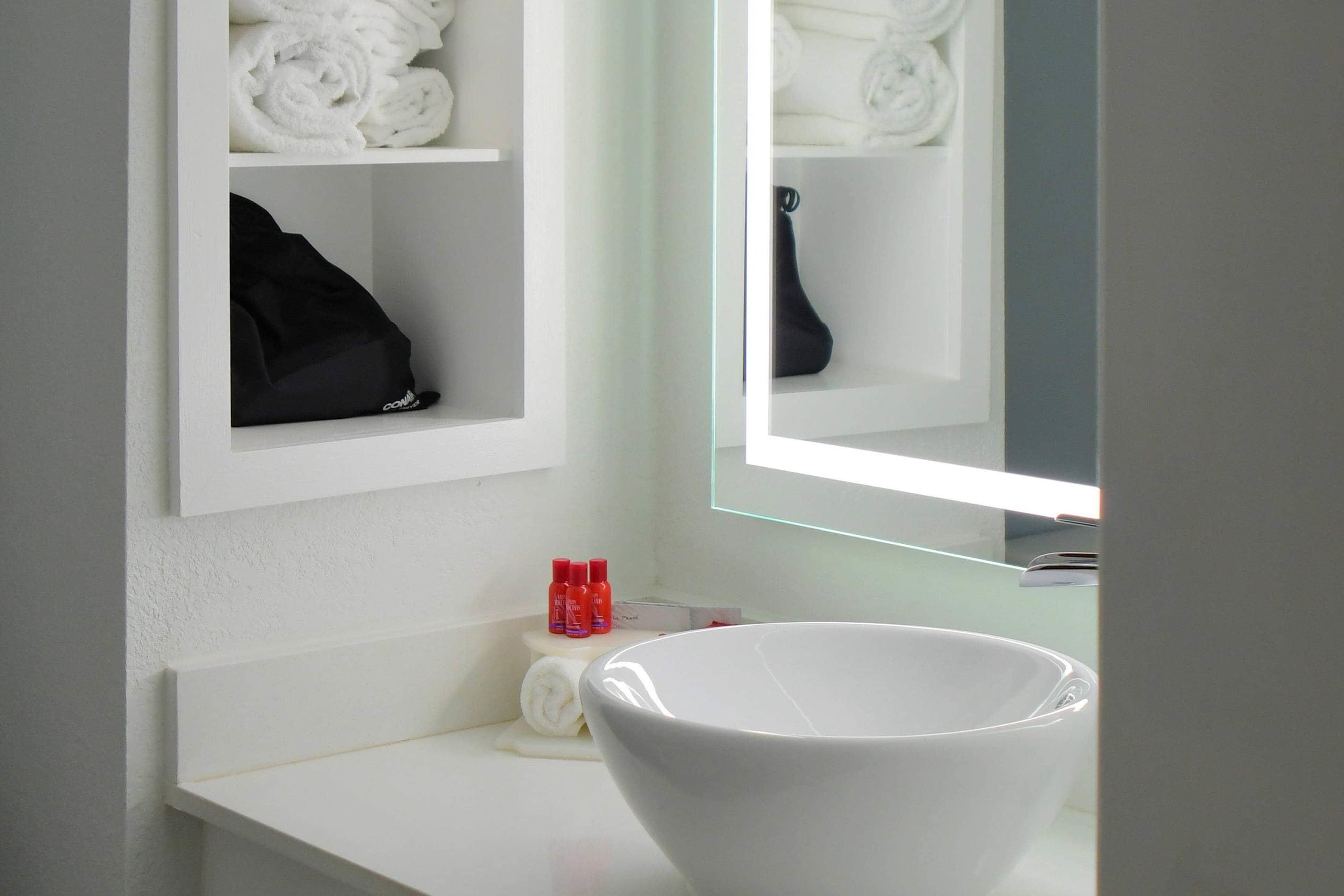A bathroom with a white sink, a mirror, and shelves with towels and toiletries.