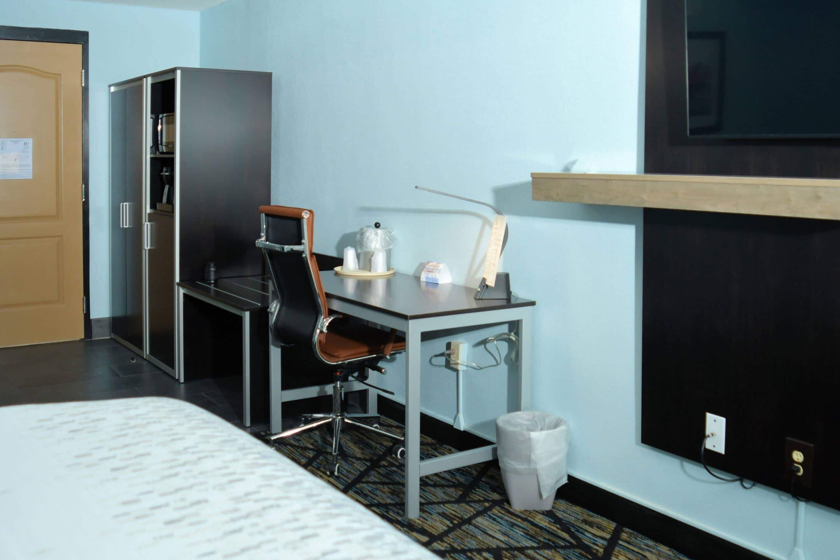 A hotel room with a bed, a desk with a chair, a small table with a lamp, a trash bin, and a wardrobe.