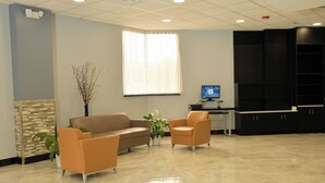 A reception area with a beige tiled floor, a beige sofa, two armchairs, a potted plant, a large window with sheer curtains, a computer monitor, and a black cabinet.