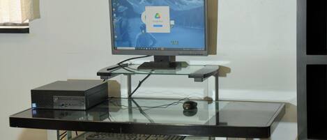 A Lenovo computer monitor on a glass desk with a keyboard and a box, possibly a server or storage unit, in a room with a white wall and a black shelf.