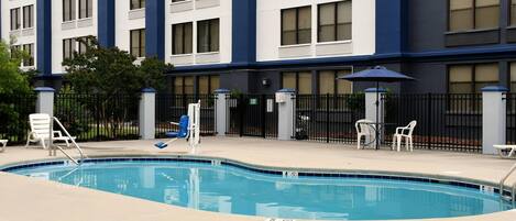 A hotel exterior with a swimming pool, outdoor seating, and a gated entrance.
