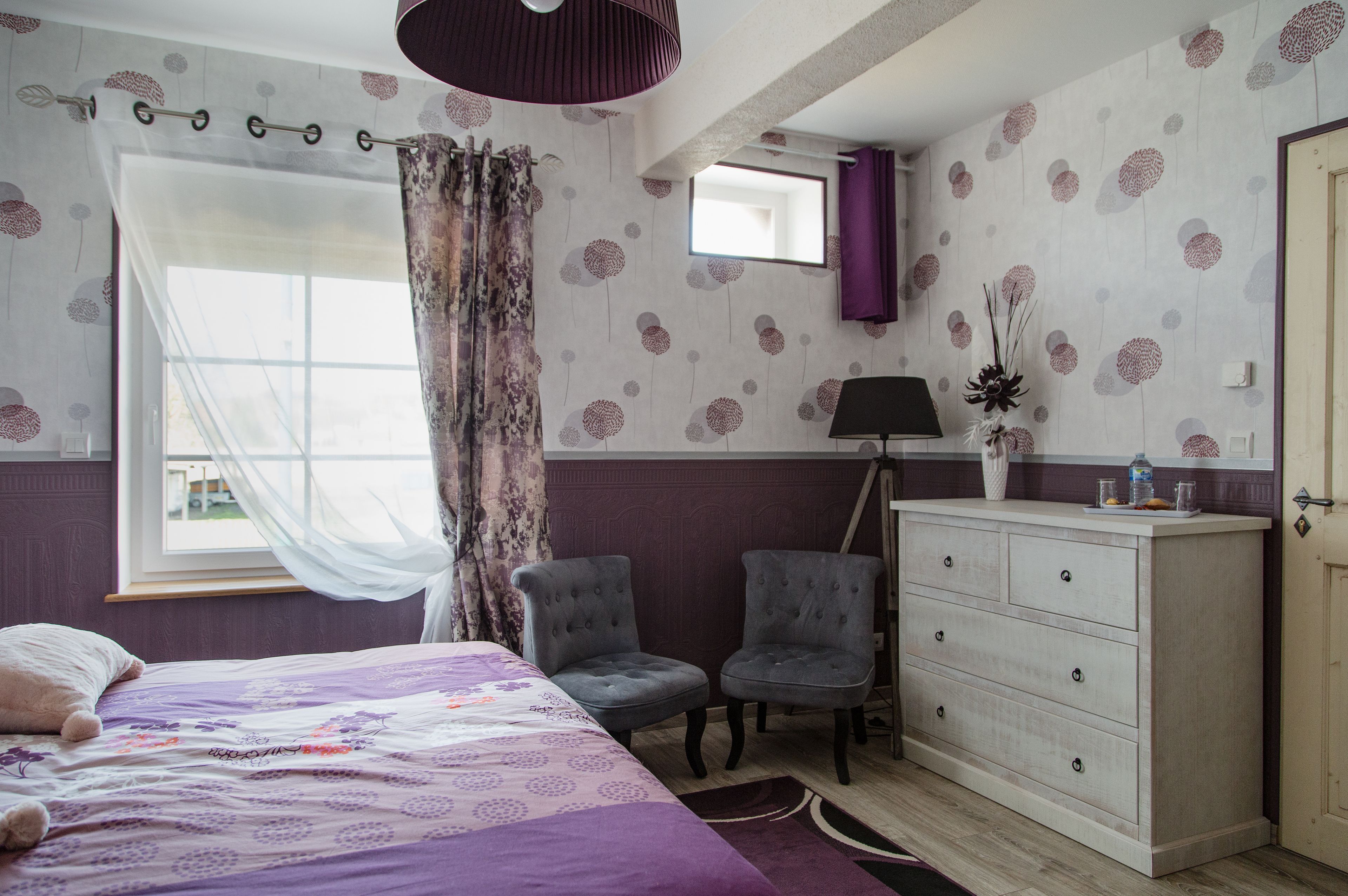 A bedroom with a bed, two chairs, a dresser, a mirror, and a window with curtains.