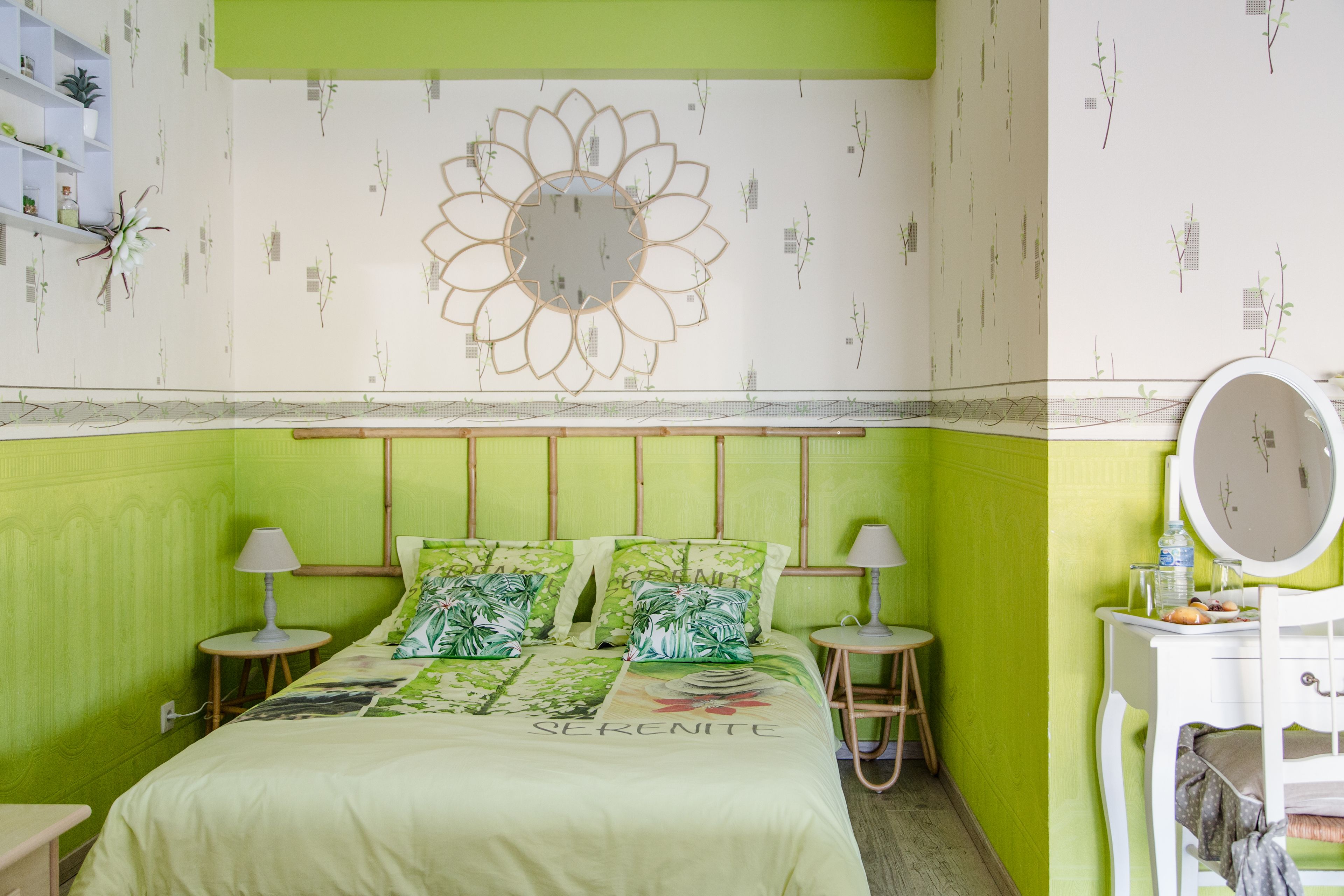 Quarto com parede verde, cama com colcha floral, duas mesas de cabeceira com luminárias, cômoda com espelho e um enfeite na parede.