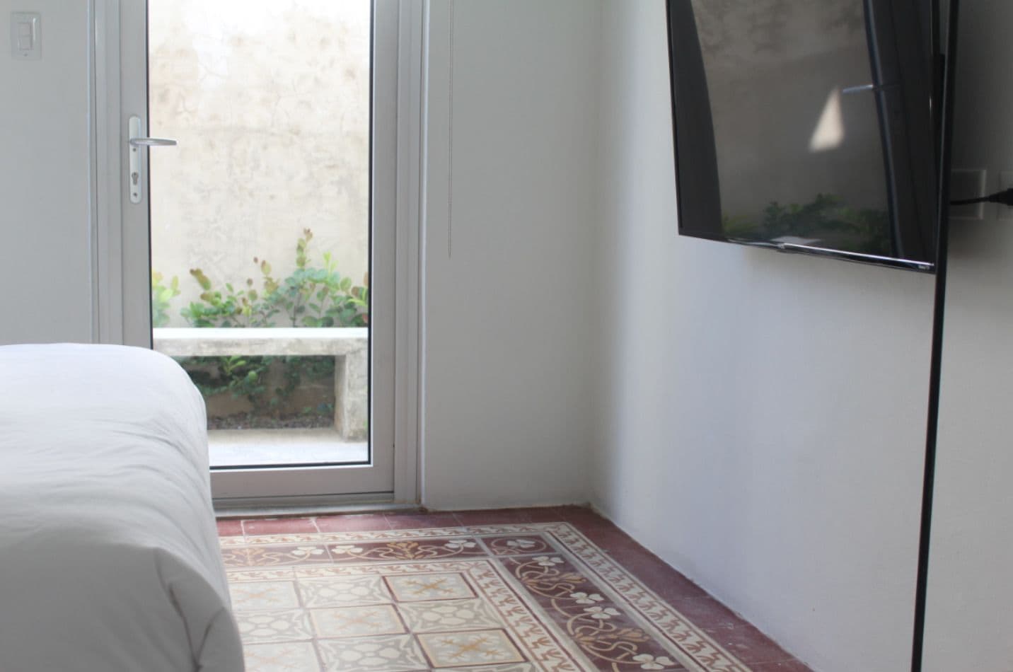 Une chambre avec un lit, un téléviseur fixé au mur et une porte-fenêtre coulissante donnant sur un espace extérieur agrémenté de plantes.