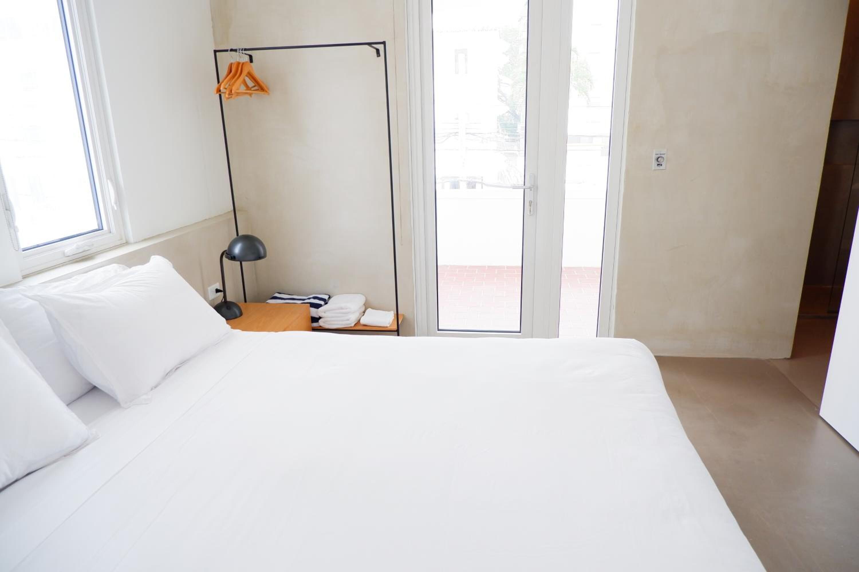 A modern hotel room with a large bed, a bedside table with a lamp, a hanging rack with clothes, and a view through the glass door.