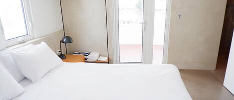 A modern hotel room with a large bed, a bedside table with a lamp, a hanging rack with clothes, and a view through the glass door.
