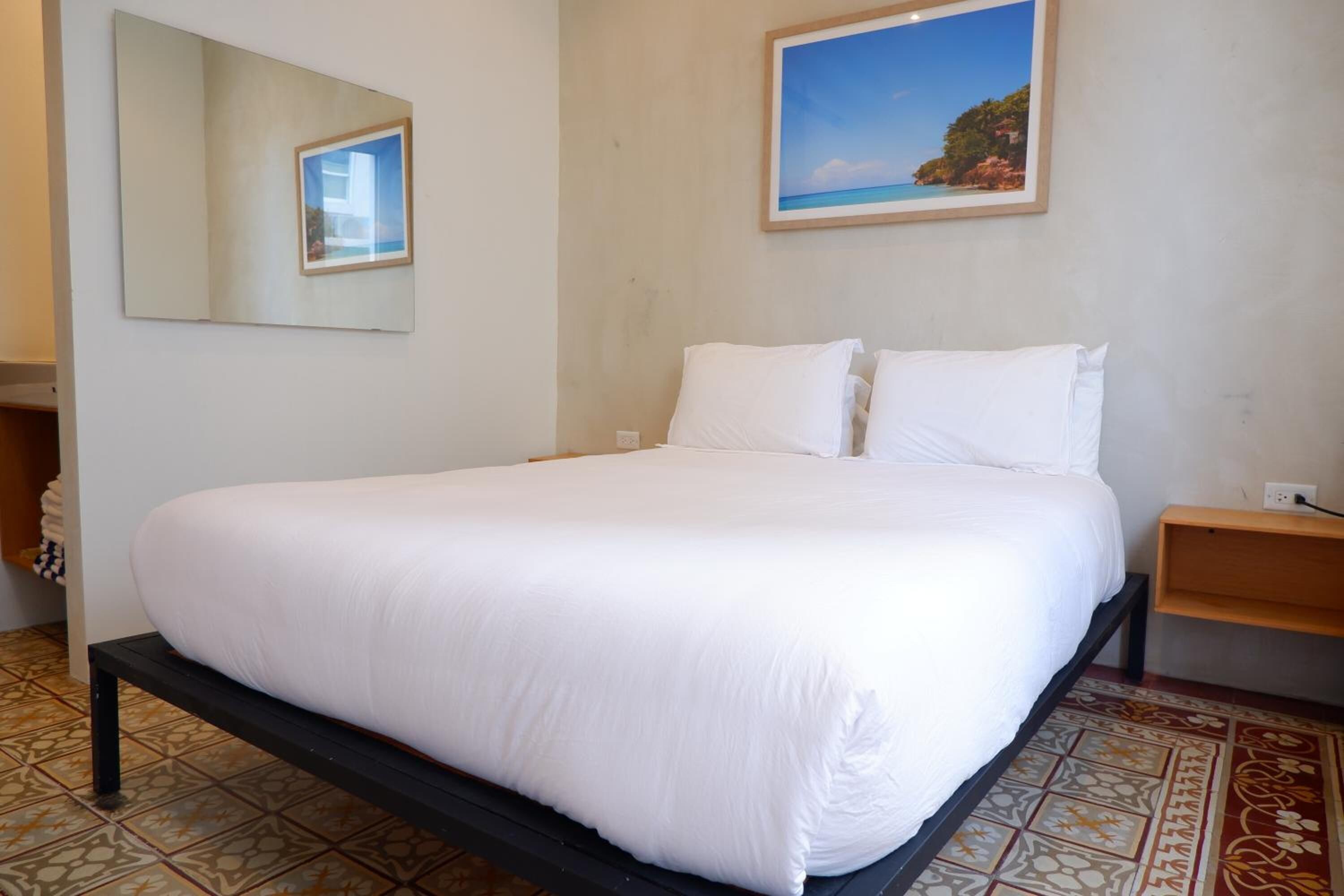 Una cama bien hecha con sábanas blancas en una habitación con piso de baldosas y un cuadro de una escena costera en la pared.