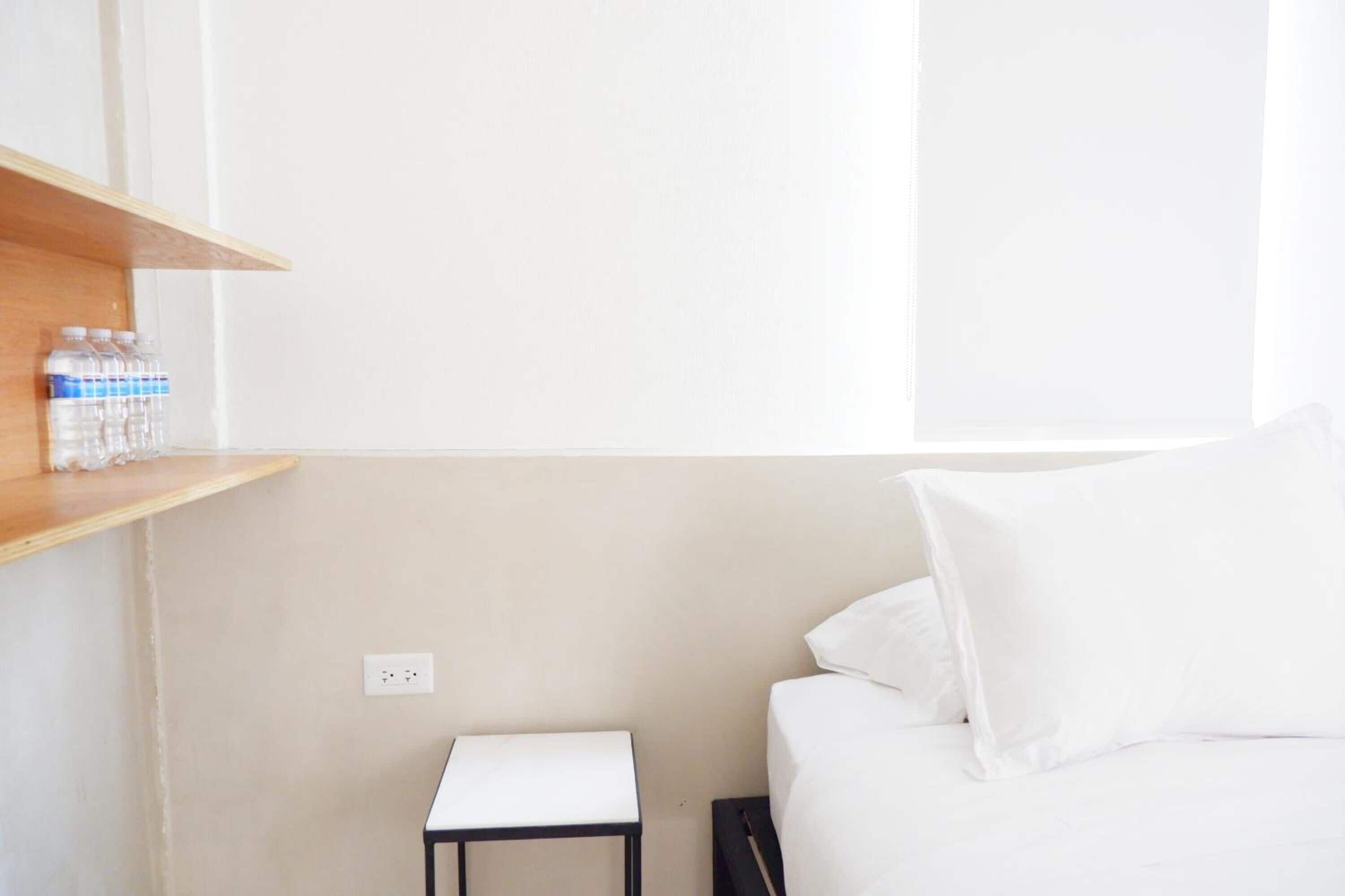 A hotel room with a bed, a small white nightstand, a shelf with water bottles, and a power outlet.