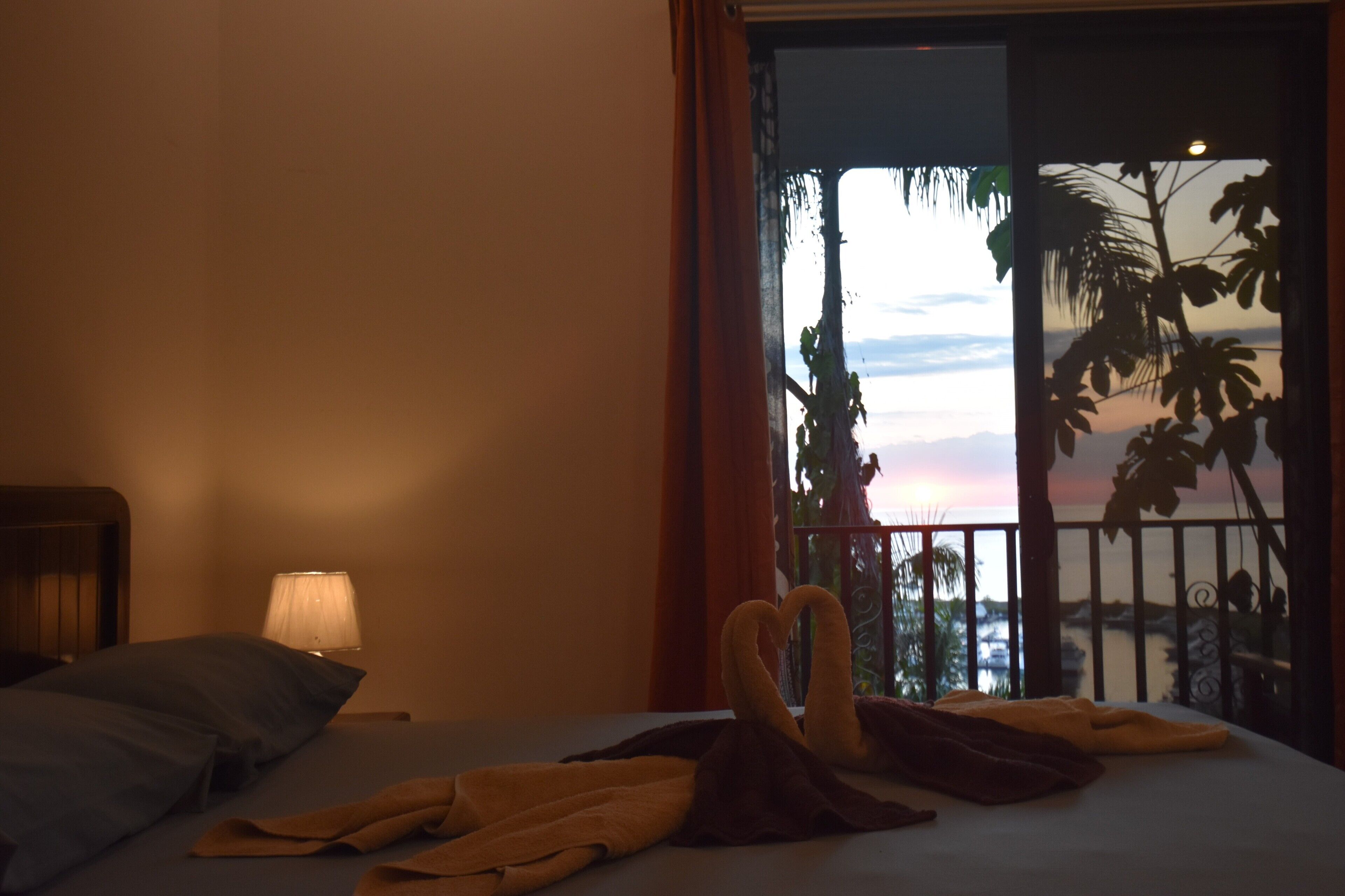 A hotel room with a bed, pillows, a heart-shaped towel arrangement, a lamp, and a balcony with a view of the sunset and palm trees.