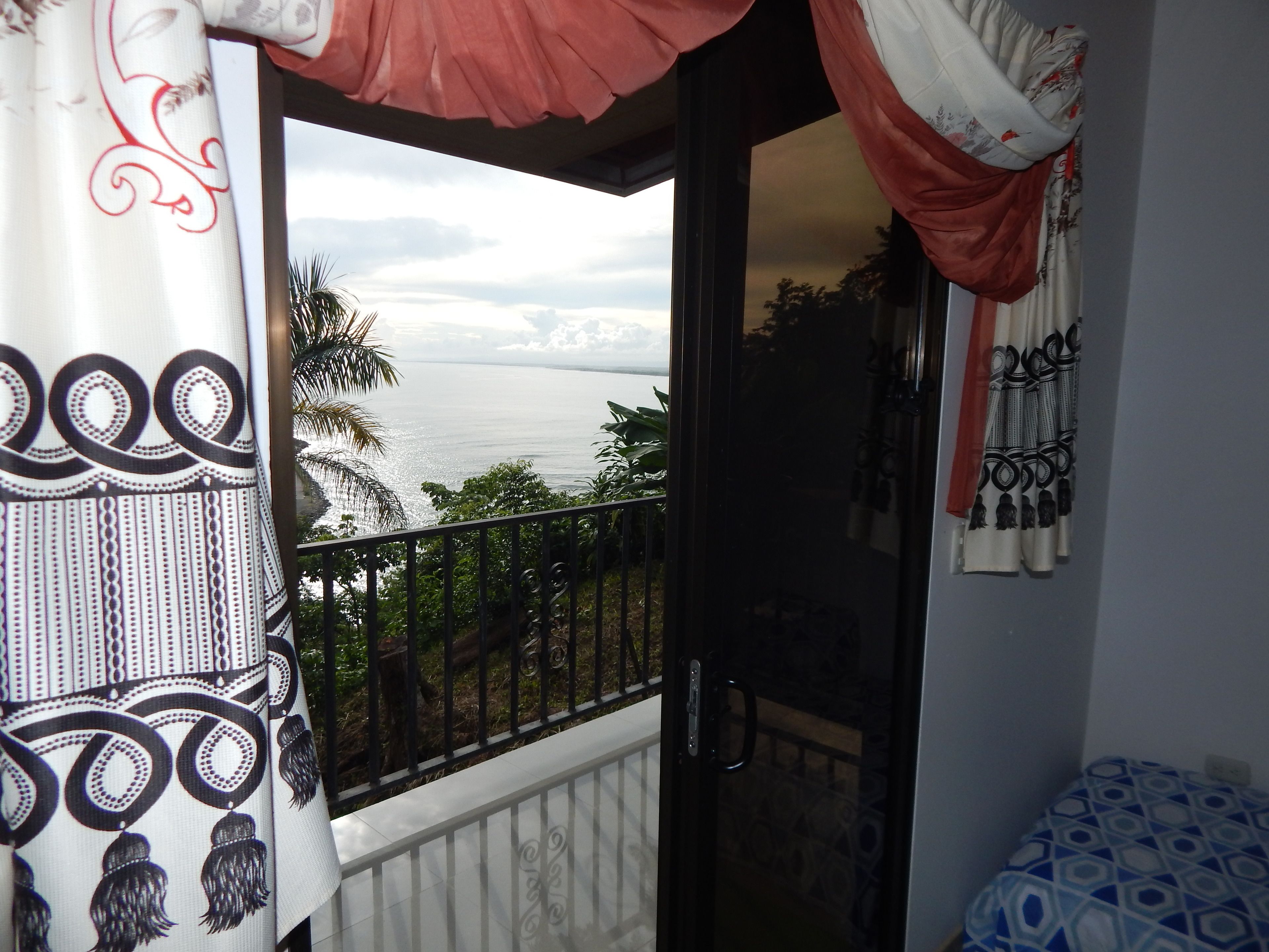 A balcony with a view of the sea, a patterned curtain, and a bed with a geometric design.