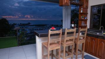 A balcony with a view of the marina and sunset. There are wooden chairs and a small table with a flower centerpiece. A bar area with a sink and shelves is visible.