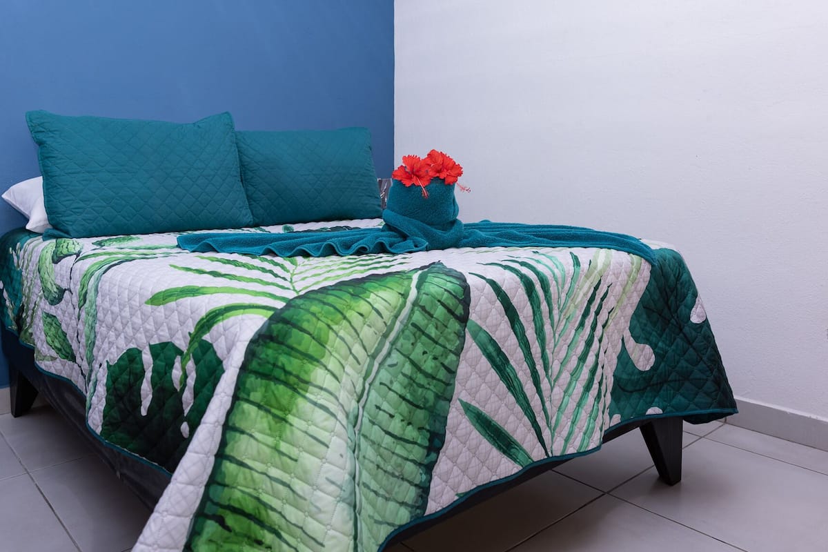 Un lit avec une couvre-lit à motifs de feuilles vertes, des coussins turquoise et un vase rempli de fleurs rouges.