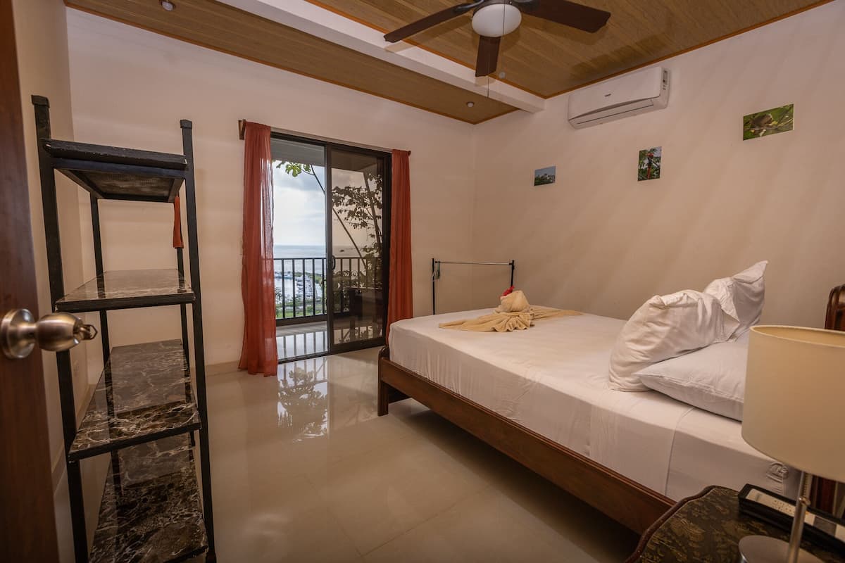 Une chambre avec un lit, un ventilateur de plafond en bois, un appareil de climatisation et une vue sur l’extérieur, visible à travers une porte vitrée.