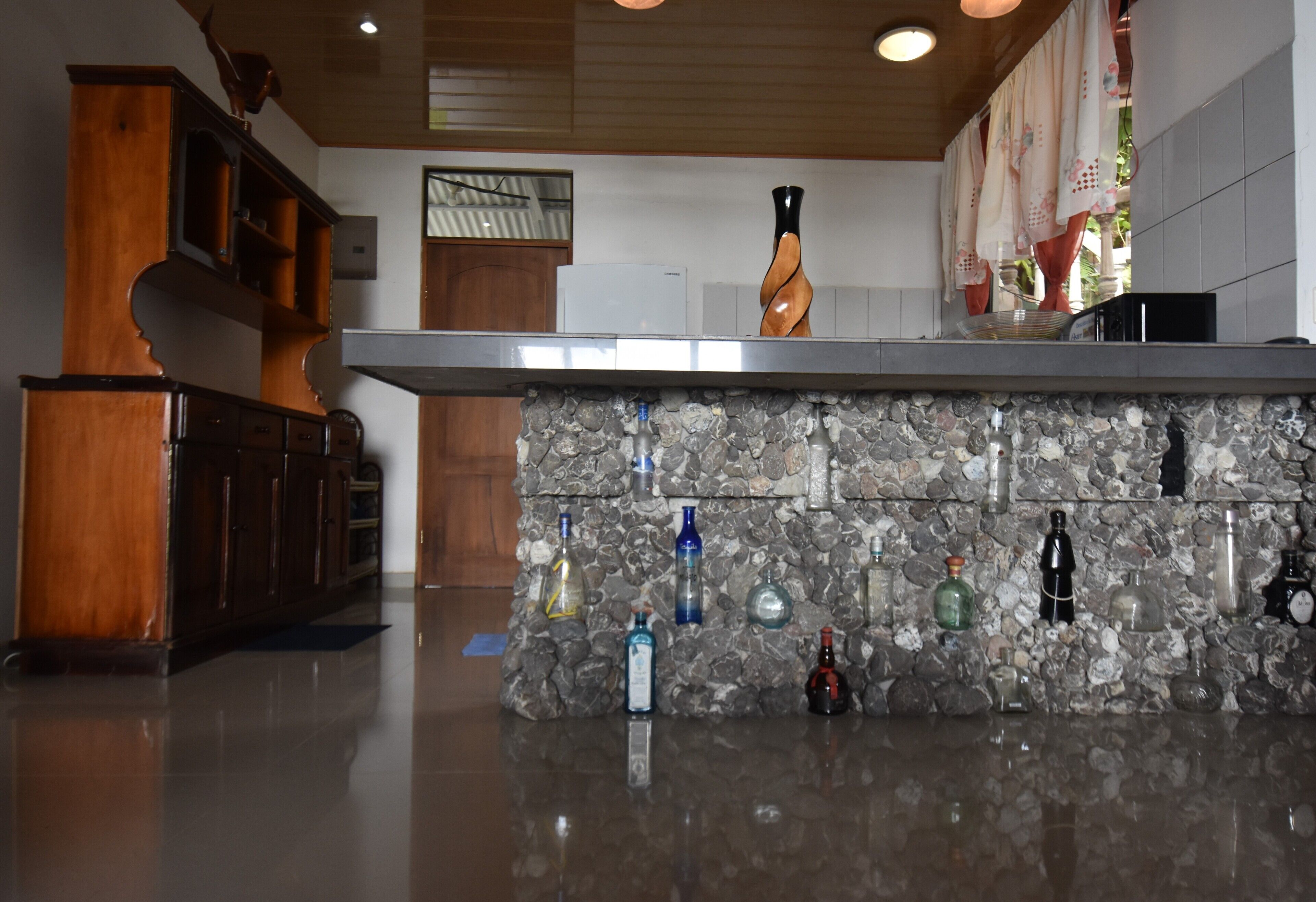 A kitchen with a stone countertop and various bottles. There is a wooden cabinet and a refrigerator.