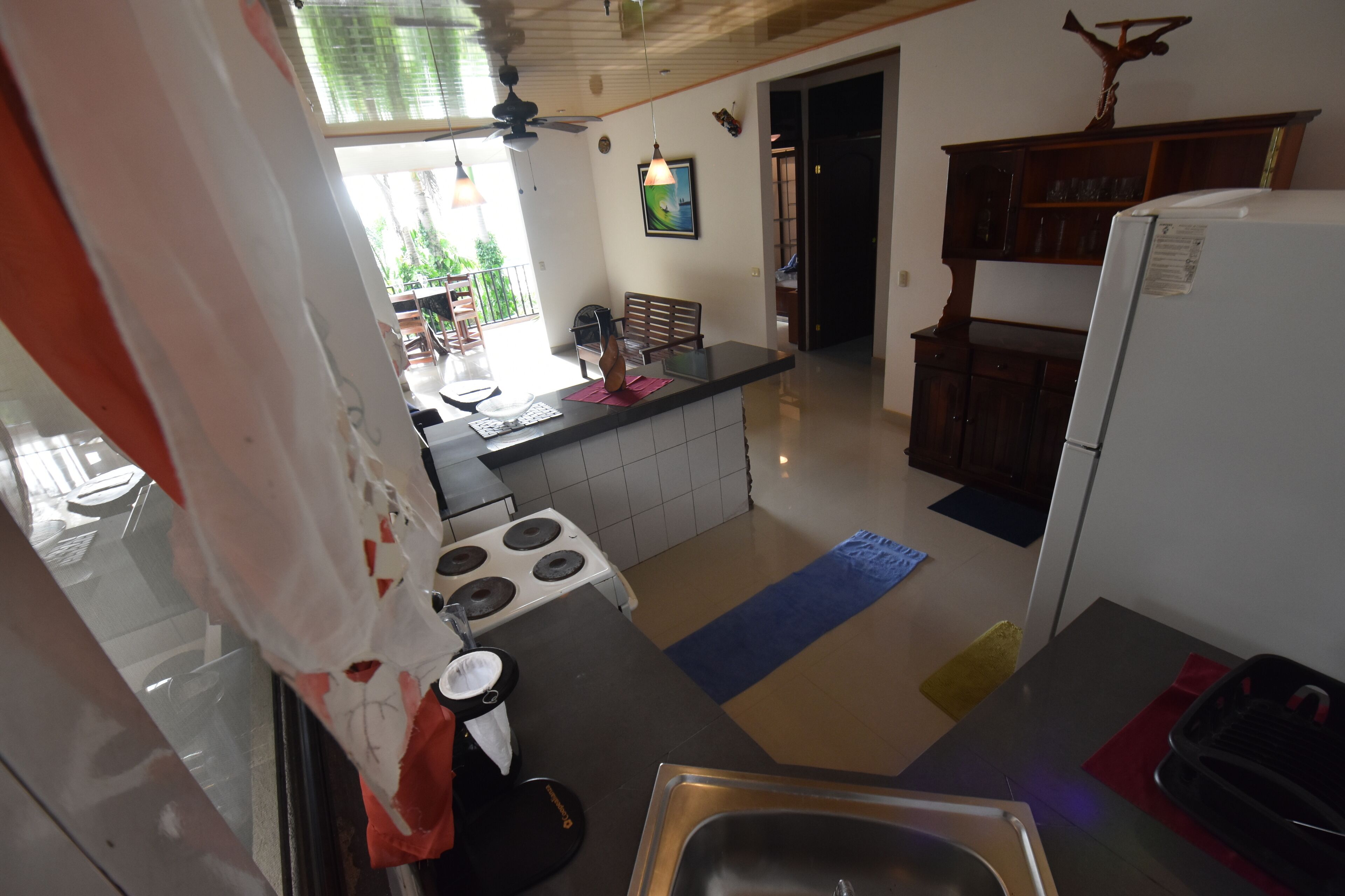 A kitchen with a stove, sink, and refrigerator. There is a window with curtains, a ceiling fan, and a wooden cabinet.