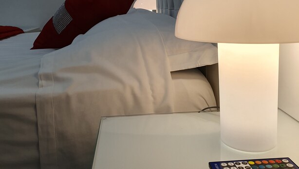 A neatly made bed with white bedding and a red pillow, a white bedside table with a remote control, and a white lamp with a white shade. - Fantasia B&B (Reggio Calabria)