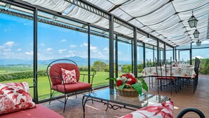 Une terrasse couverte avec un mur de verre, une chaise rouge, une table avec un panier de fleurs et un coin repas avec une table dressée pour deux personnes.