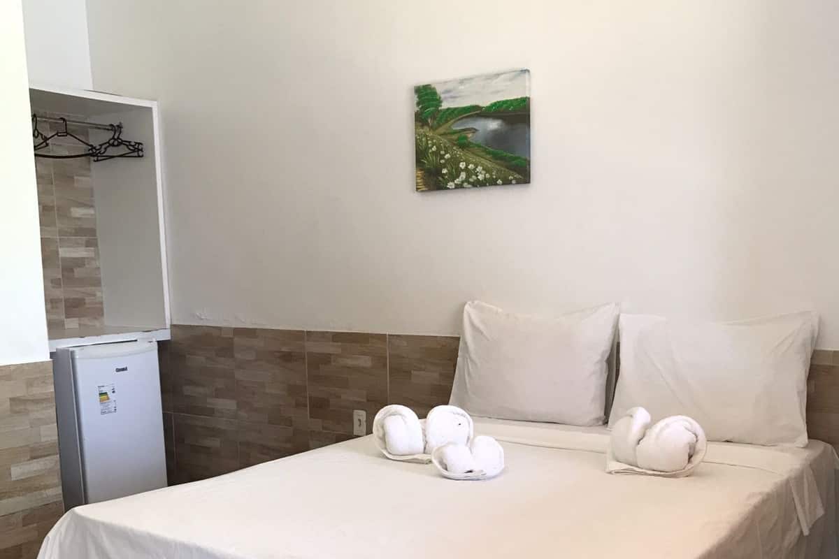 Una habitación de hotel con una cama, almohadas blancas, un refrigerador pequeño y un cuadro en la pared.