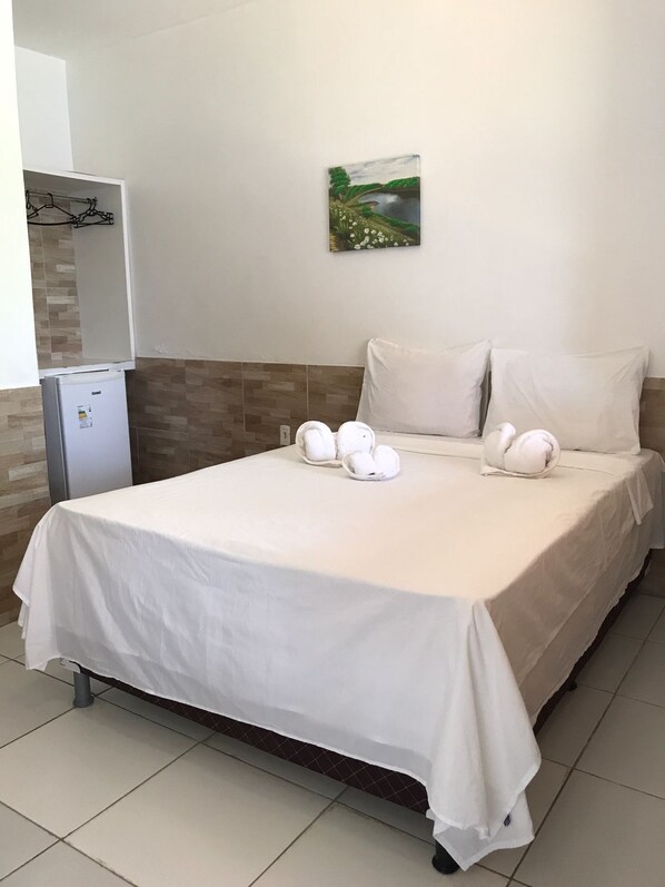 A hotel room with a bed, white pillows, a small refrigerator, and a painting on the wall. - Pousada Recanto das Arvores (Barreirinhas)