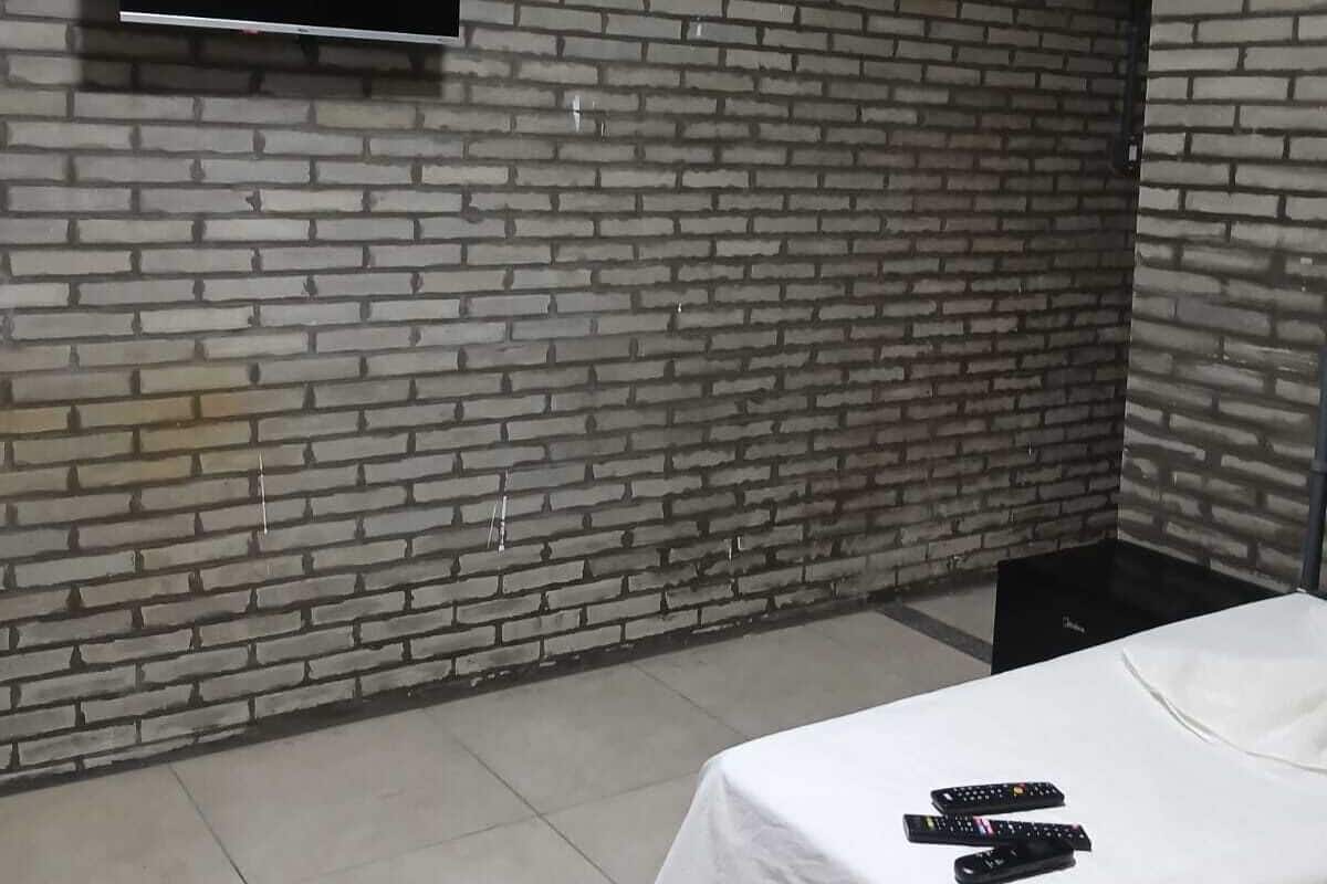 Una habitación con pared de ladrillo, un televisor de pantalla plana sobre la cama y varios controles remotos sobre la misma.