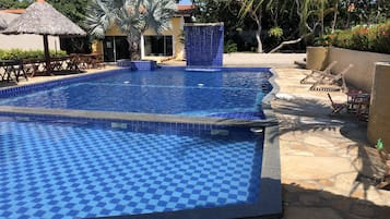 Una piscina con diseño de tablero de ajedrez, rodeada de sillas de descanso y una palmera.