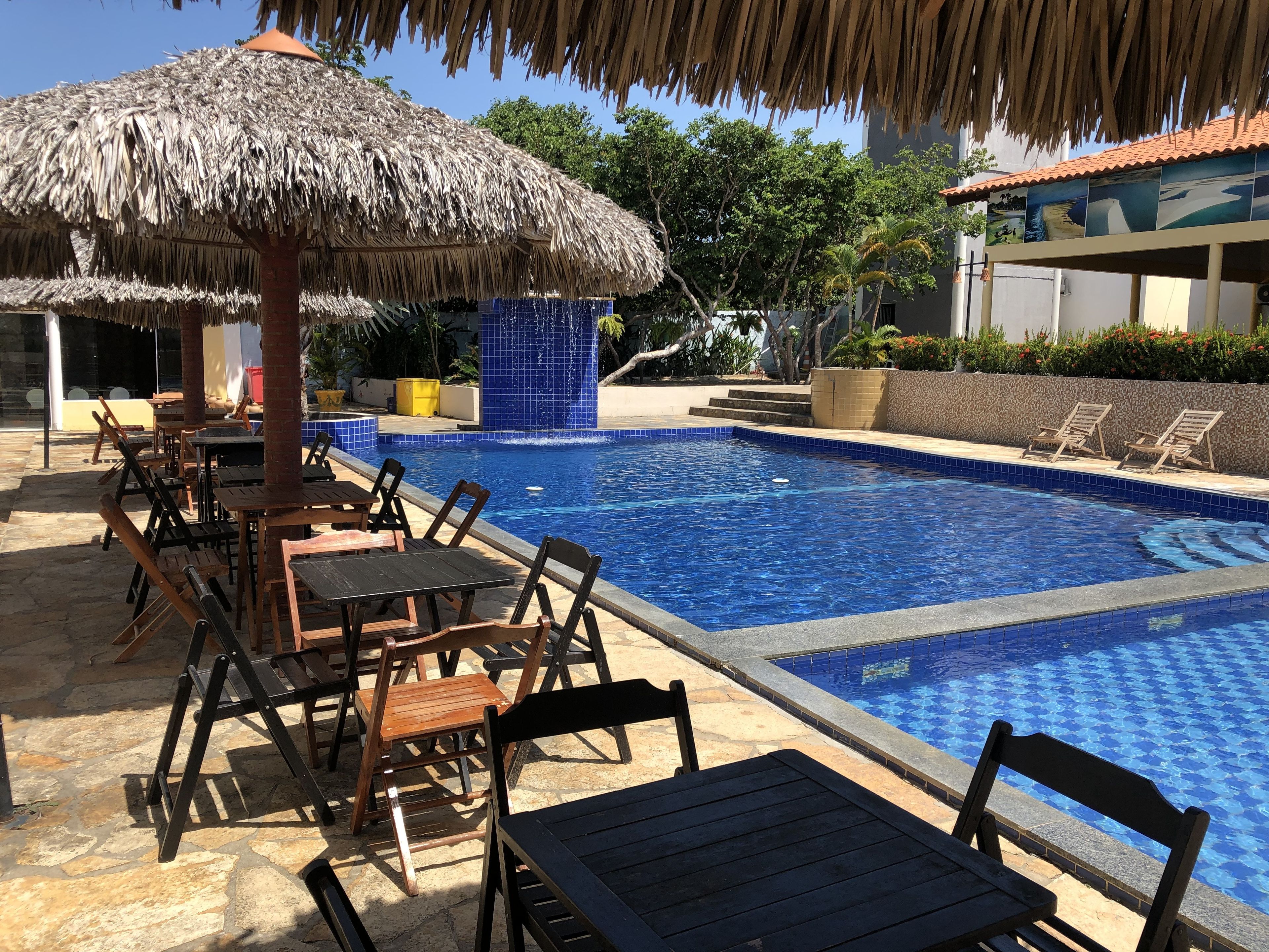 Una piscina con sillas y mesas de descanso, una sombrilla de paja y una pared de azulejos con una cascada.