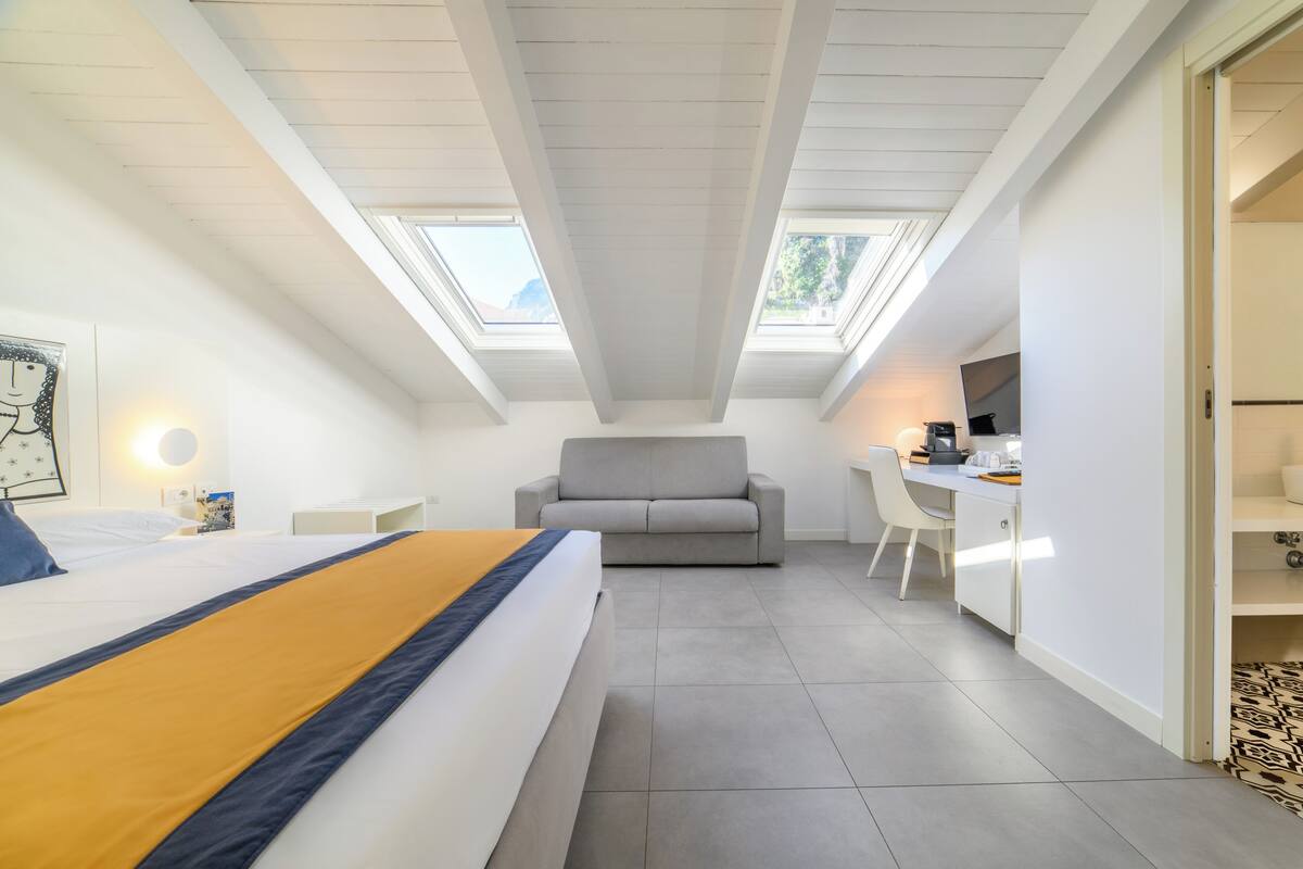 Un dormitorio moderno con tragaluz, una cama con colcha amarilla y azul, un sofá gris, un escritorio y una silla.