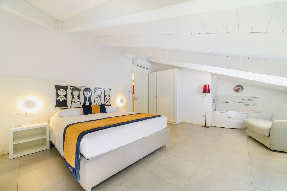 Un dormitorio moderno con una cama grande, mesitas de noche, una lámpara de pie roja, un sofá blanco y obras de arte en las paredes.