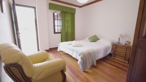 Quarto de hotel com cama, poltrona, cômoda e janela com cortinas verdes.