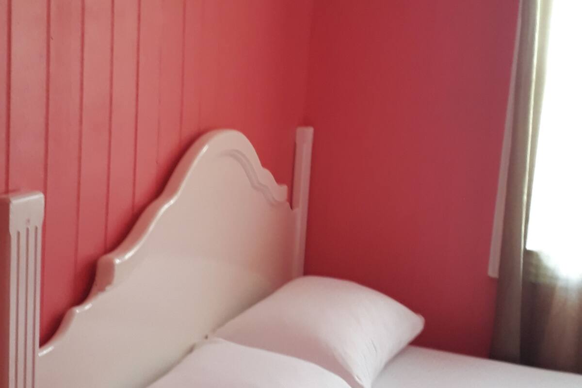 Una habitación con una cama frente a una pared roja, un cabecero blanco y una ventana con cortinas.