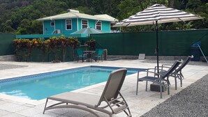 Una piscina con sillas de descanso y un paraguas a rayas, rodeada por una cerca verde y una casa al fondo.