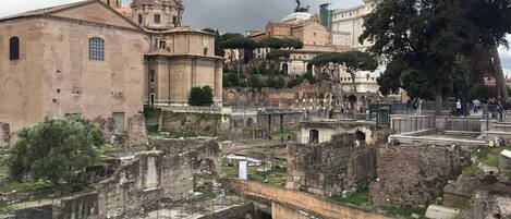 Un antiguo sitio romano con ruinas, incluyendo una gran cúpula y estatuas, rodeado de árboles y edificios modernos.