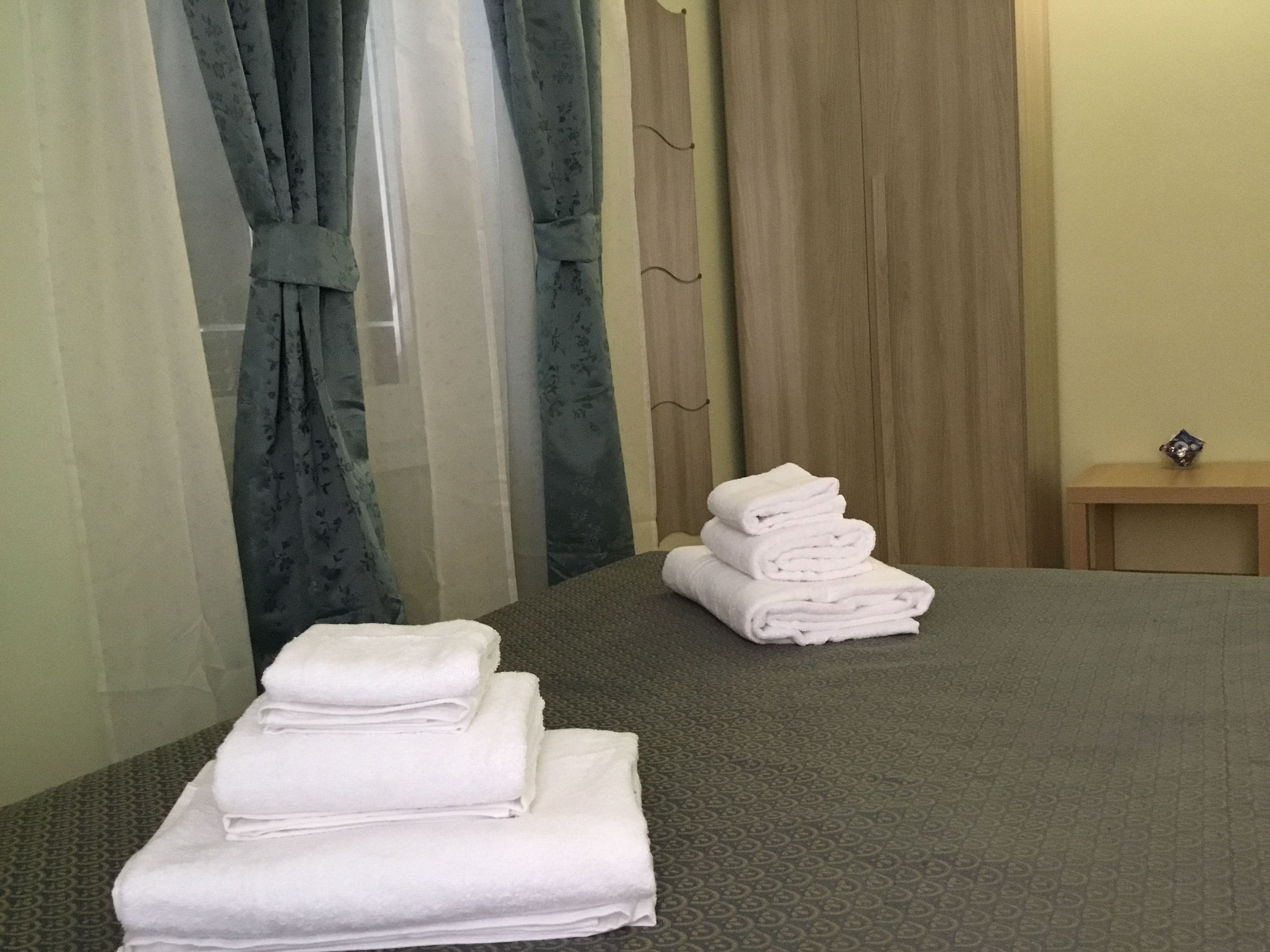 A hotel room with a bed, a stack of white towels, a wooden nightstand, and a small table with a decorative item.