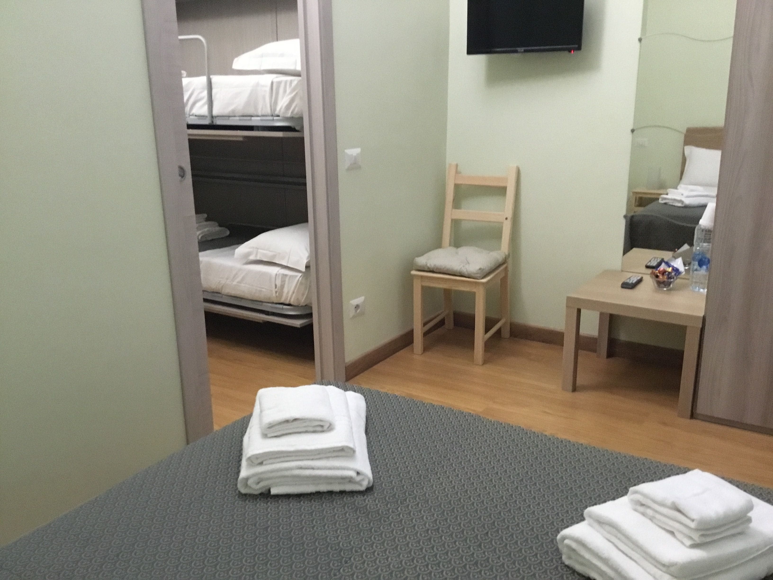 A hotel room with a bunk bed, a single bed, a chair, a small table, and a TV mounted on the wall.