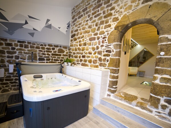 A modern bathroom with a stone wall, a hot tub, and a wooden archway leading to another room. - La Cazette (Severac-d'Aveyron)