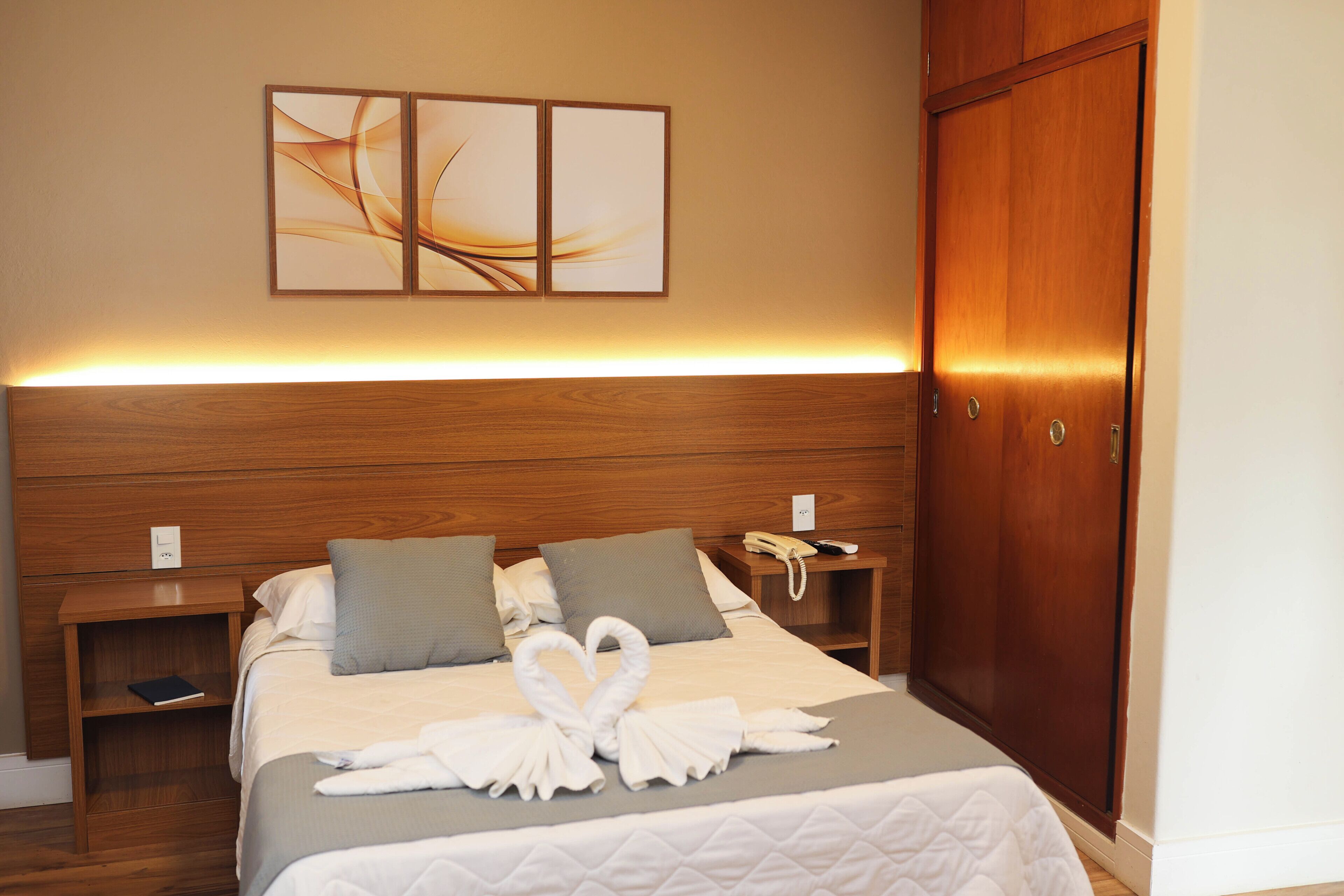 Une chambre d’hôtel avec une tête de lit en bois, un lit recouvert d’une courtepointe matelassée, deux oreillers, une table de chevet avec un livre, un téléphone et une œuvre d’art accrochée au mur.