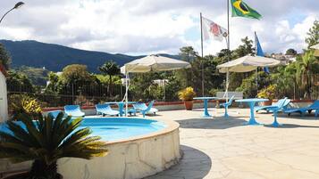 Zona de piscina con sillas, sombrillas, banderas y montañas al fondo.