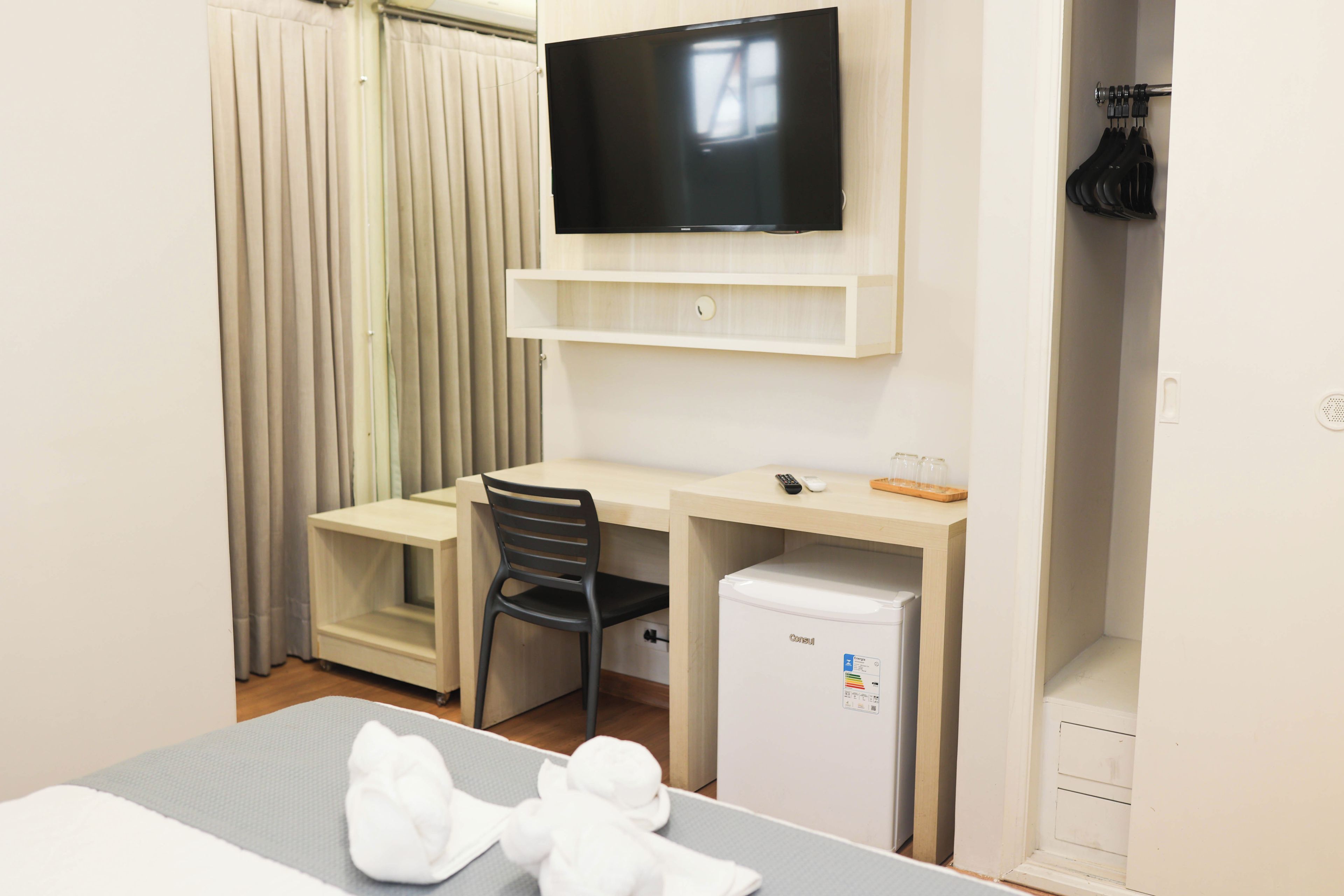Habitación de hotel con cama, escritorio con silla, televisor montado en la pared, refrigerador pequeño y armario con ropa colgada.