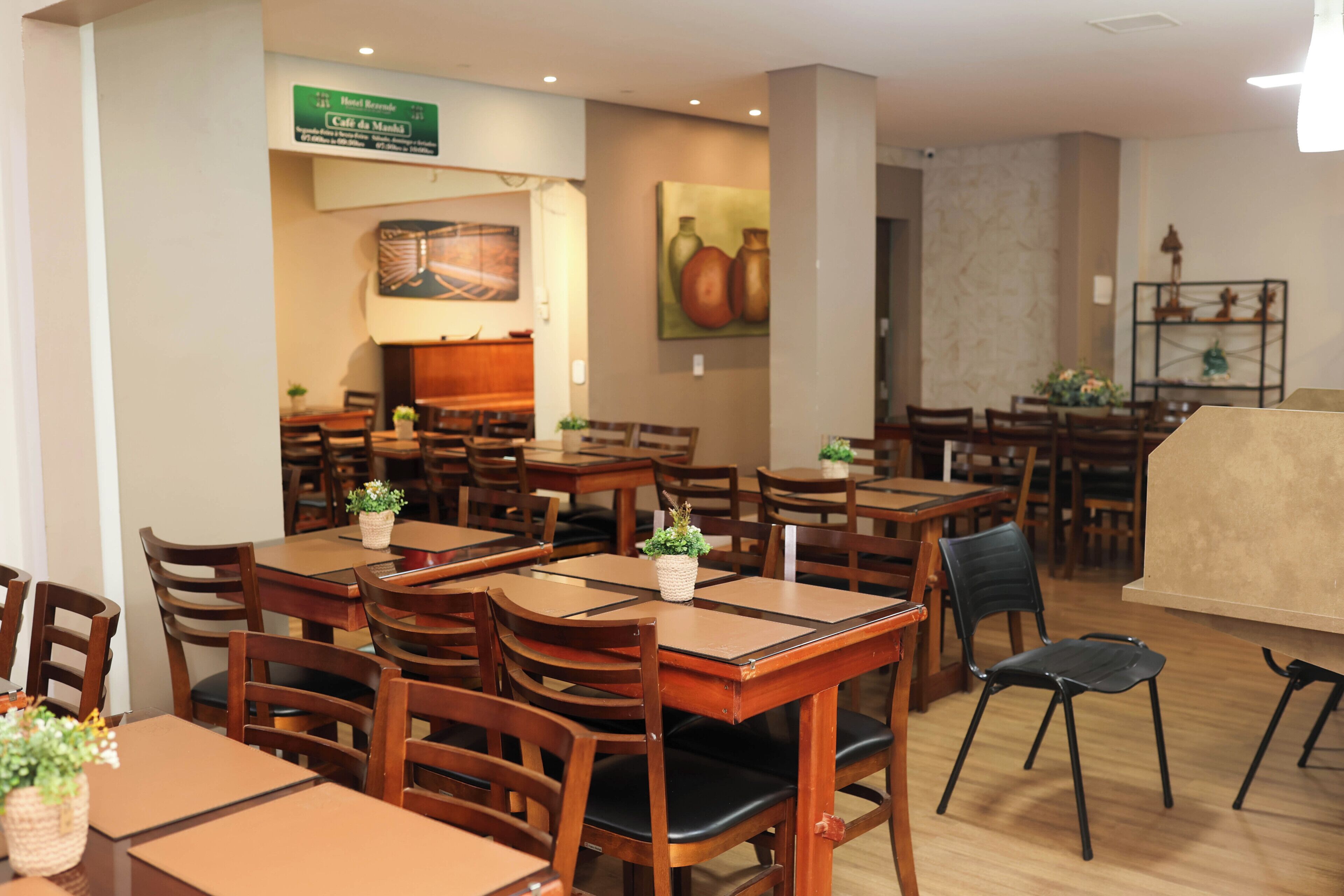 Un comedor de hotel con varias mesas y sillas de madera, un letrero verde que indica una cafetería y una pared con una pintura de frutas.