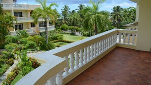 Uma varanda com balaustrada branca, com vista para um jardim ajardinado com palmeiras e um edifício com várias varandas.