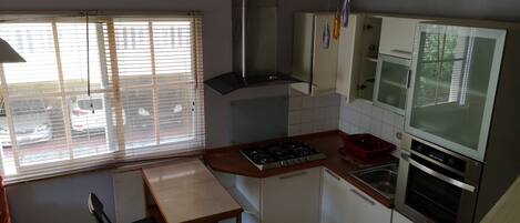 A compact kitchen with a gas stove, oven, and a microwave. There is a window with blinds, a small dining table with chairs, and a cabinet with a glass door.