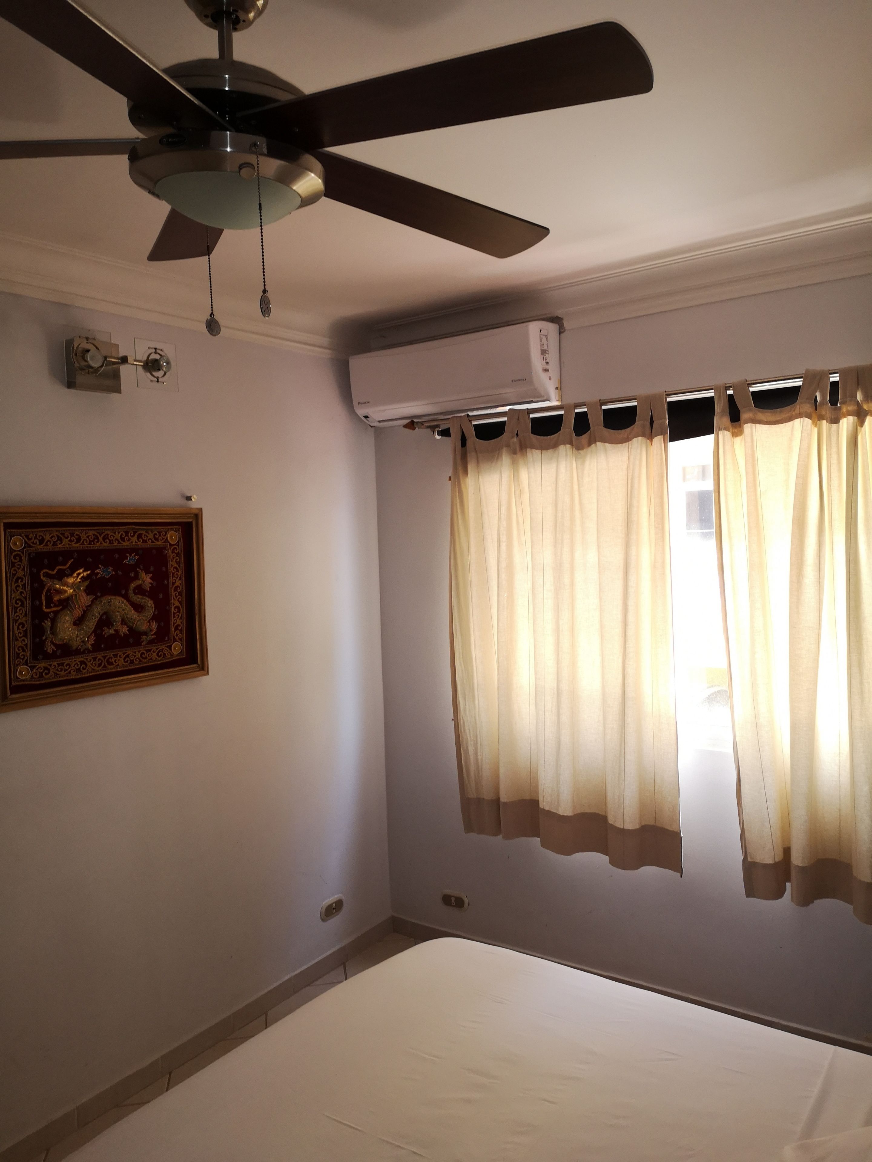 A room with a ceiling fan, an air conditioning unit, a bed, and a window with curtains.