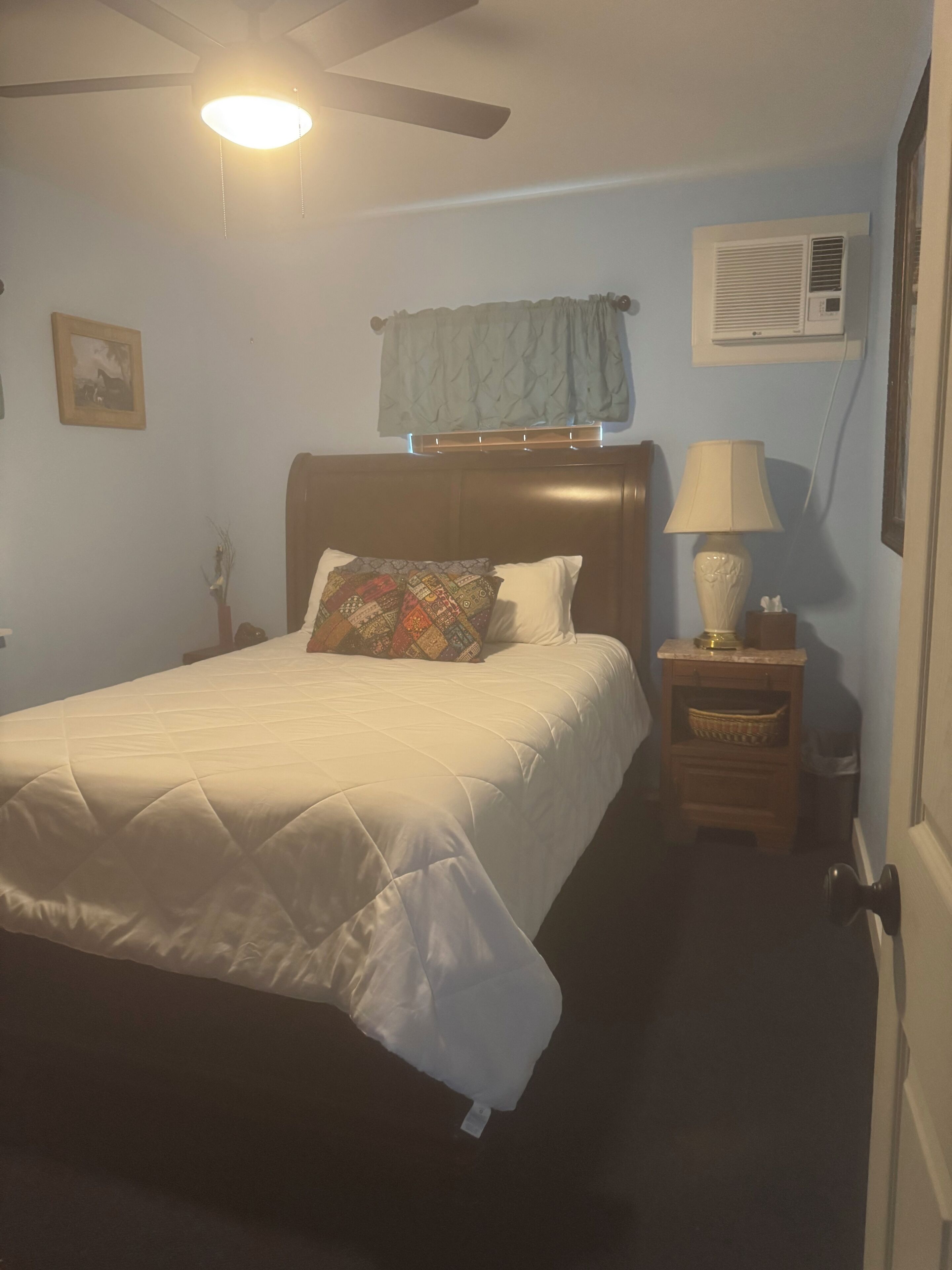 Un dormitorio con cama, mesita de noche, lámpara, aire acondicionado, ventilador de techo y ventana con cortina.
