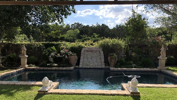 Una piscina con un muro de piedra y estatuas, rodeada de un césped cuidado y diversas plantas.