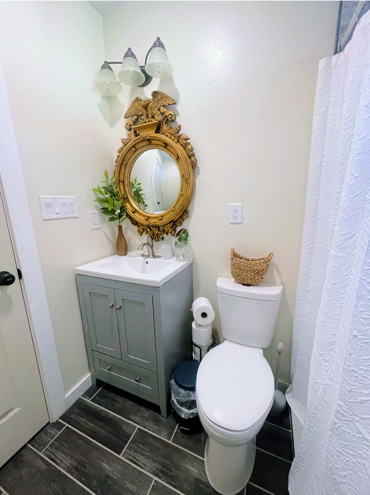 A bathroom with a white toilet, a grey cabinet, a gold-framed mirror, and a wall-mounted light fixture.