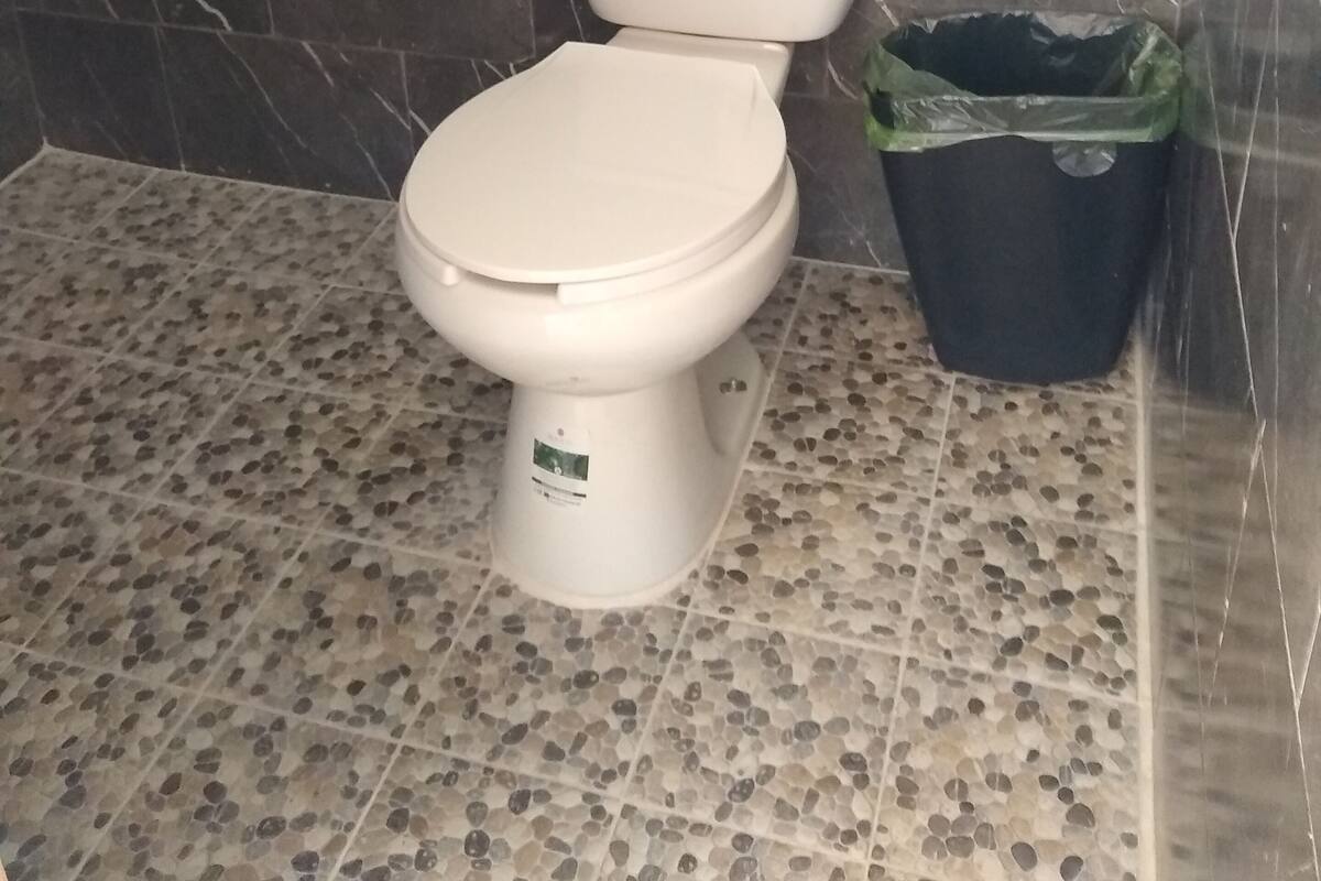 Un inodoro con asiento y tapa blancos, un cesto de basura negro con bolsa verde, y un suelo de baldosas con diseño de piedrecitas.