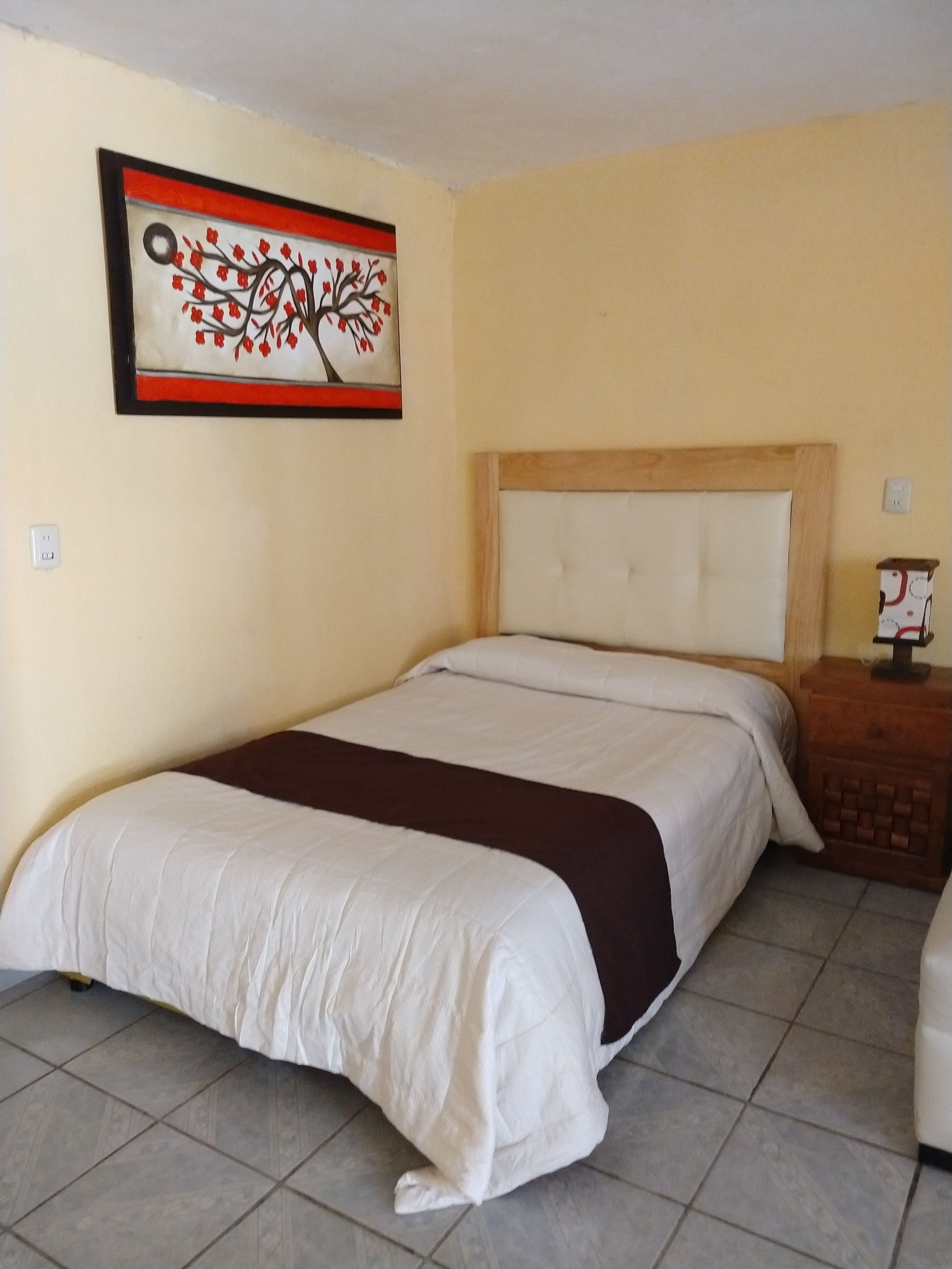 A hotel room with a large bed, a headboard, a nightstand, a wall-mounted painting, and a tiled floor.