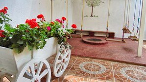 Un patio cubierto con una maceta blanca llena de flores rojas, un conjunto de columpios y un mural de un árbol en la pared.