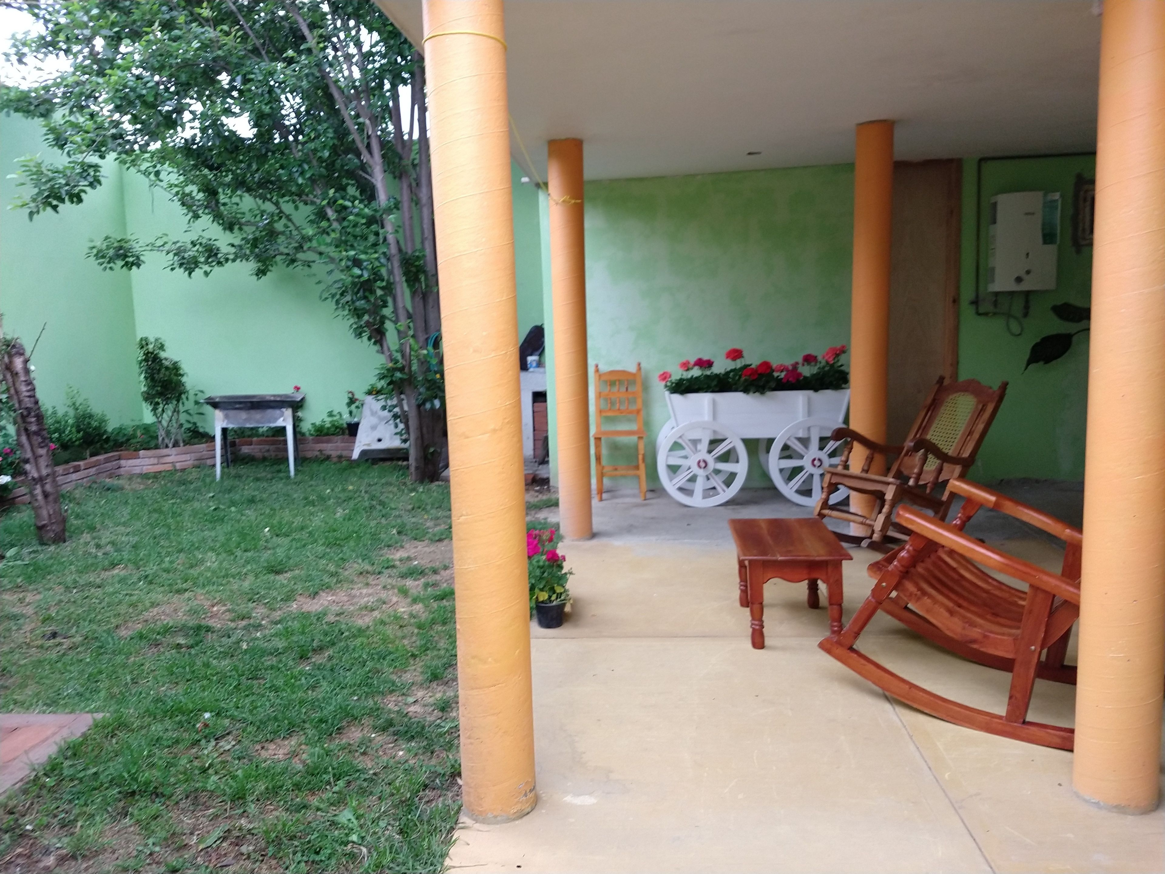 Un espace extérieur abrité comprenant un fauteuil à bascule en bois, une petite table et un chariot blanc décoré de fleurs.