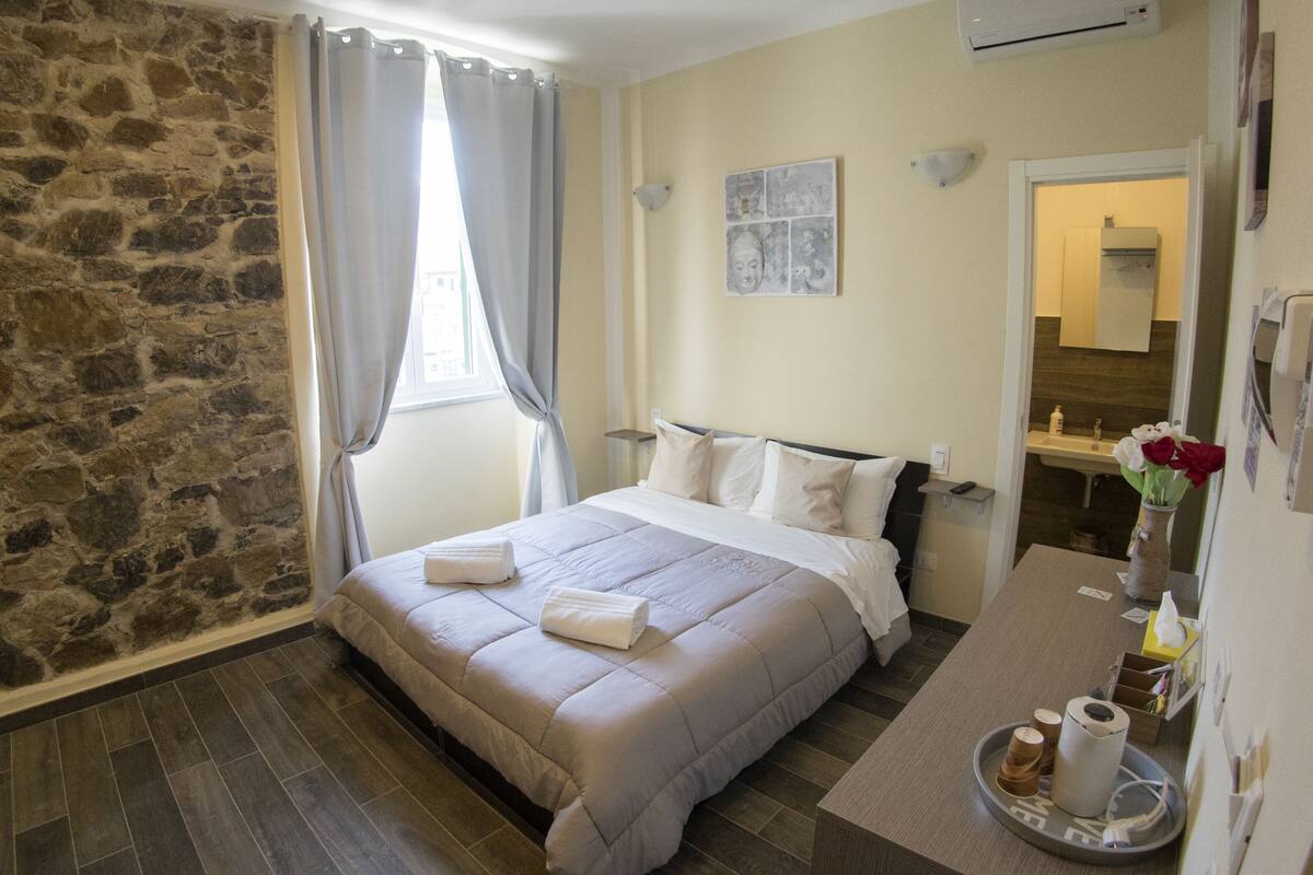 Ein Schlafzimmer mit Steinwand, einem Bett mit weißer Bettwäsche, einem Nachttisch mit Blumenvase und einem durch eine offene Tür einsehbaren Badezimmer.